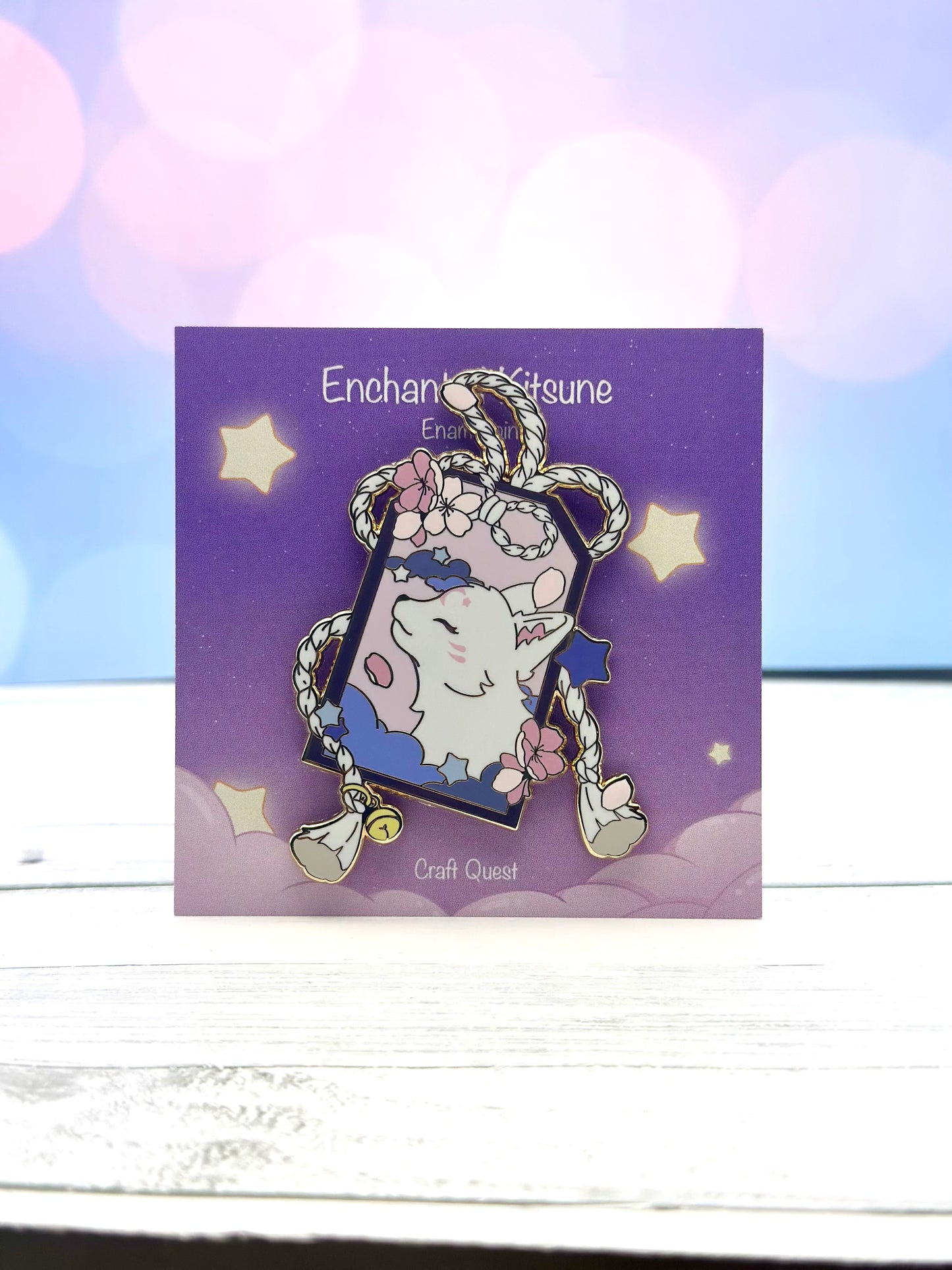 Enamel pin featuring a white kitsune framed by braided rope, blue clouds, stars, and pink blossoms on a purple display card.