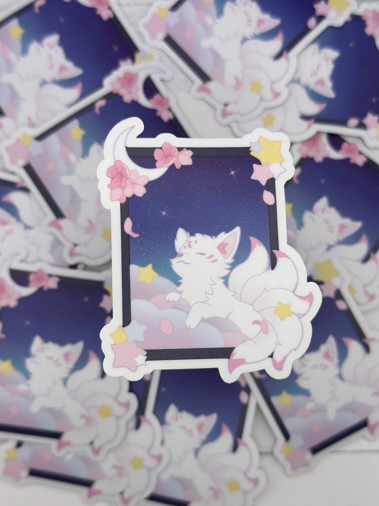 Pastel kitsune sticker featuring a white fox resting on fluffy clouds inside a moonlit window frame decorated with stars and cherry blossoms.