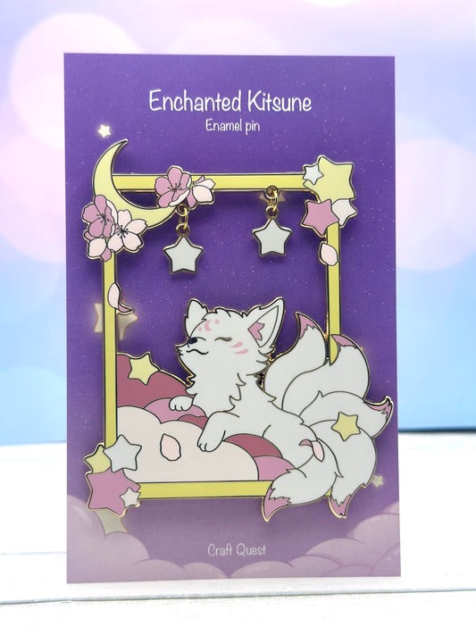 White enchanted kitsune enamel pin resting on pink clouds inside a gold frame with cherry blossoms, crescent moon, and dangling star charms on a purple backing card