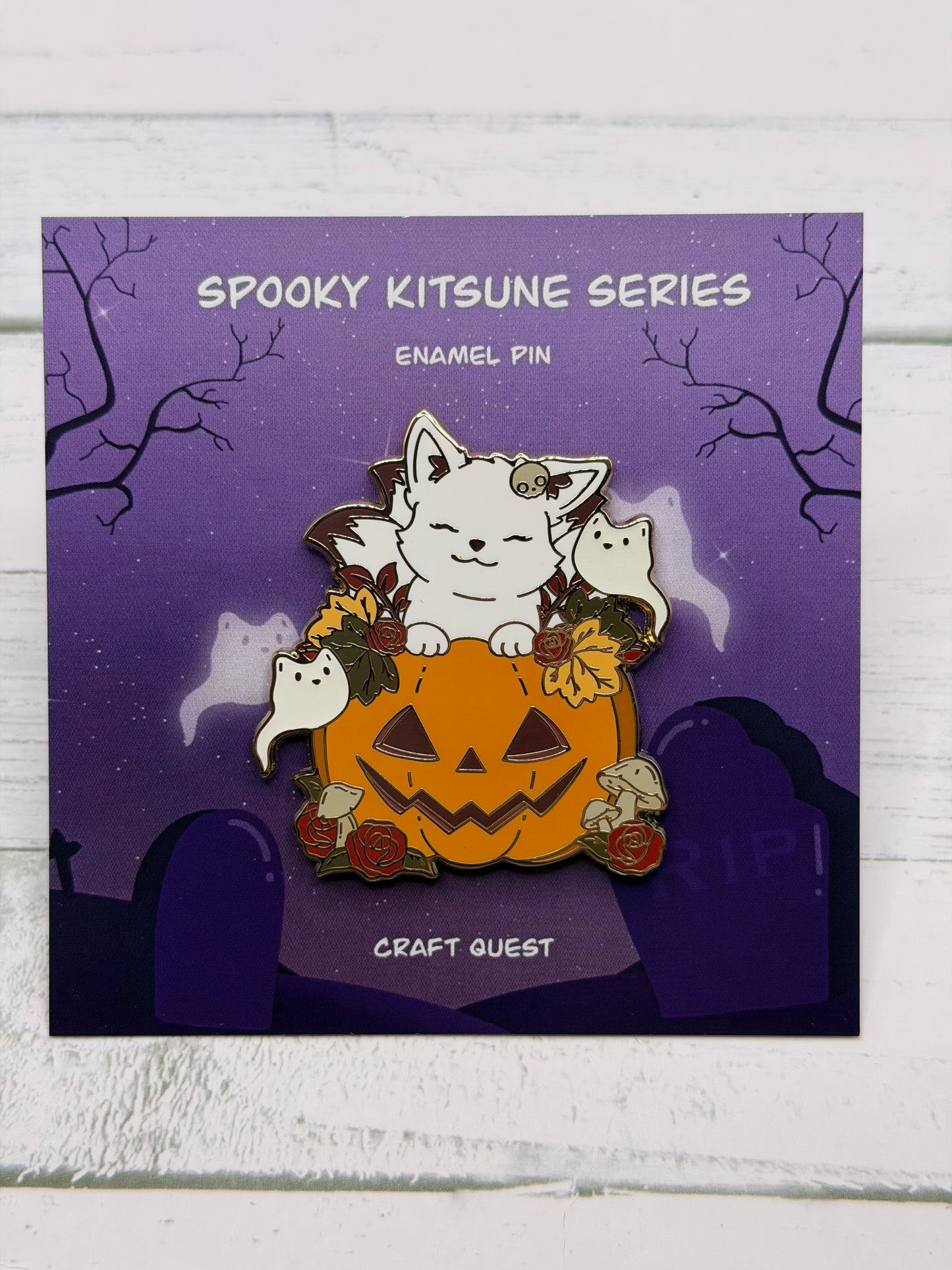 White kitsune enamel pin resting on a carved jack-o’-lantern surrounded by ghosts, roses, and mushrooms on a purple spooky graveyard backing card