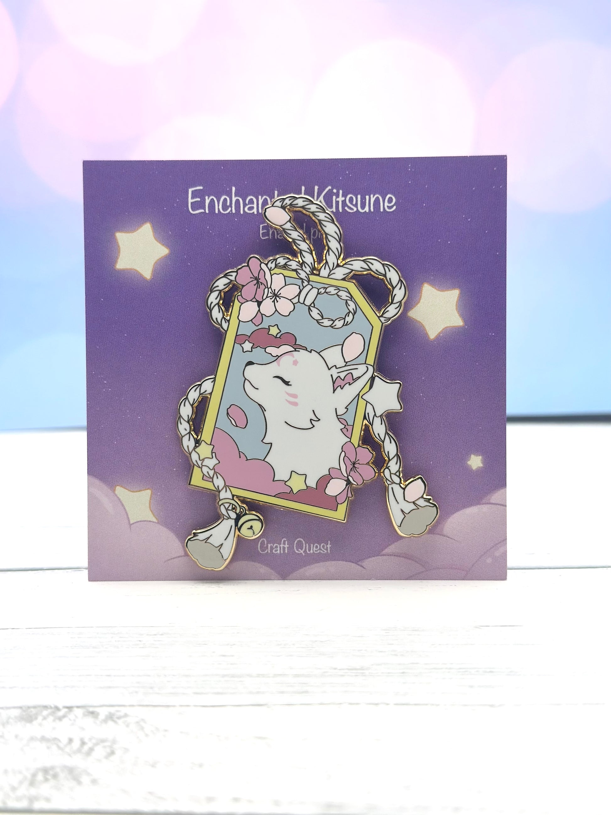 Enamel pin featuring a white kitsune framed by braided rope, pink clouds, stars, and pink blossoms on a purple display card.