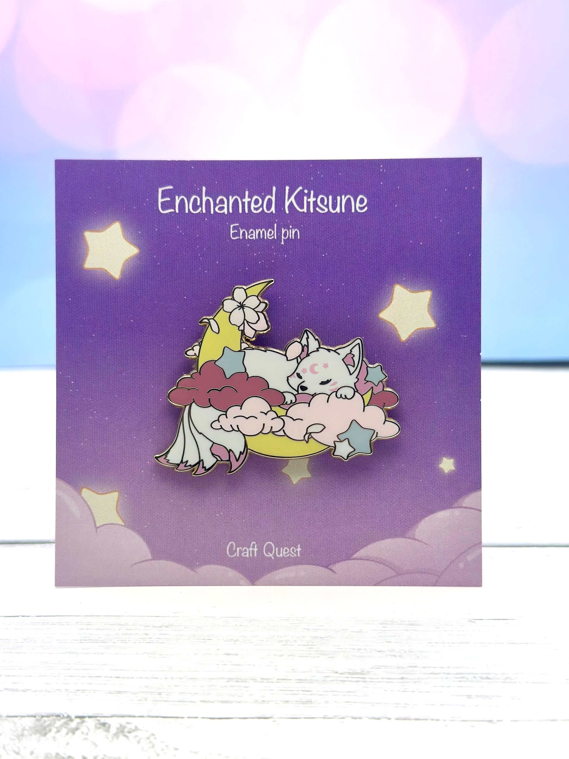 Enamel pin featuring a sleeping white kitsune resting on pink clouds and a crescent moon on a purple display card.