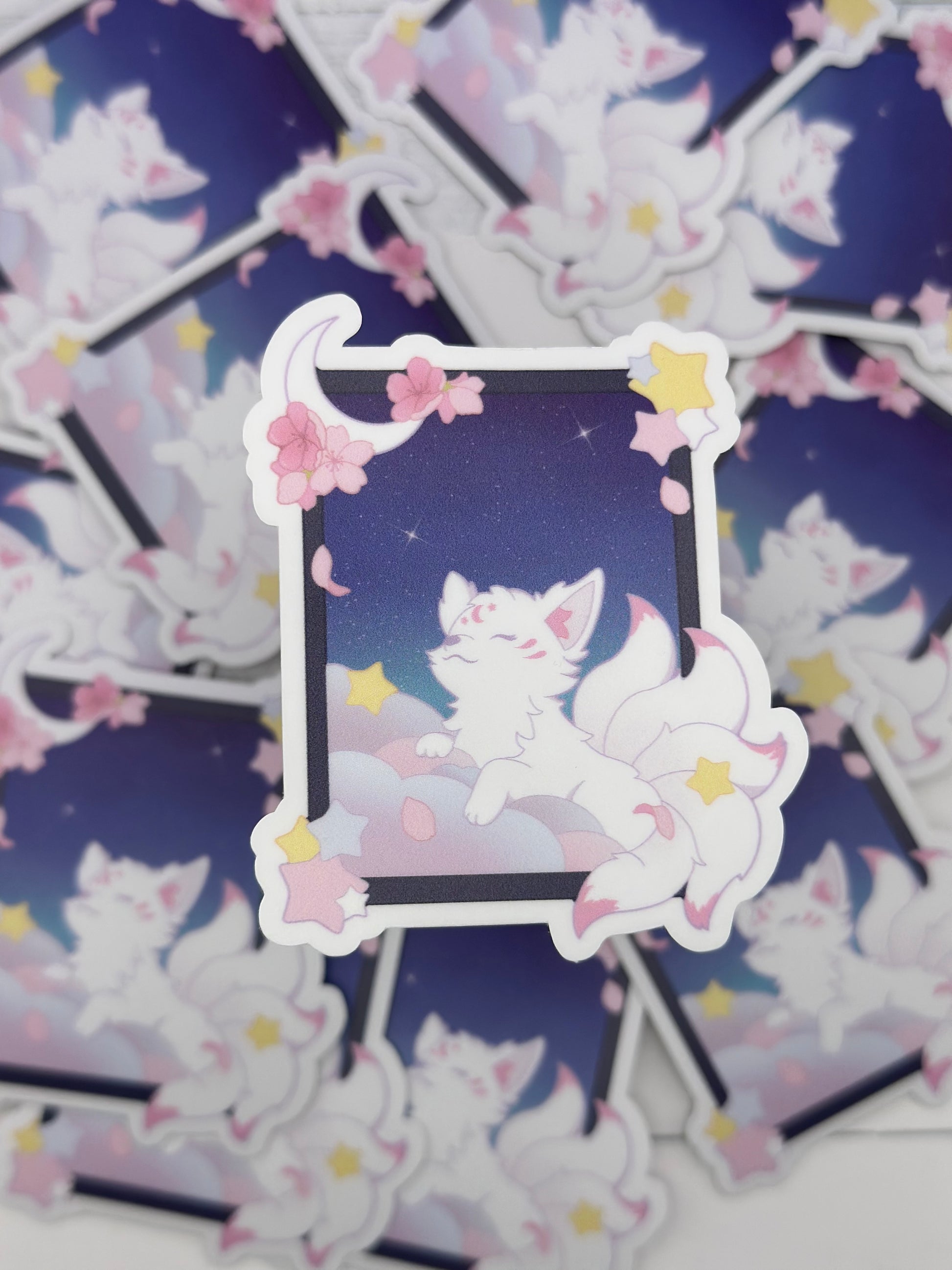 Pastel kitsune sticker featuring a white fox resting on fluffy clouds inside a moonlit window frame decorated with stars and cherry blossoms.
