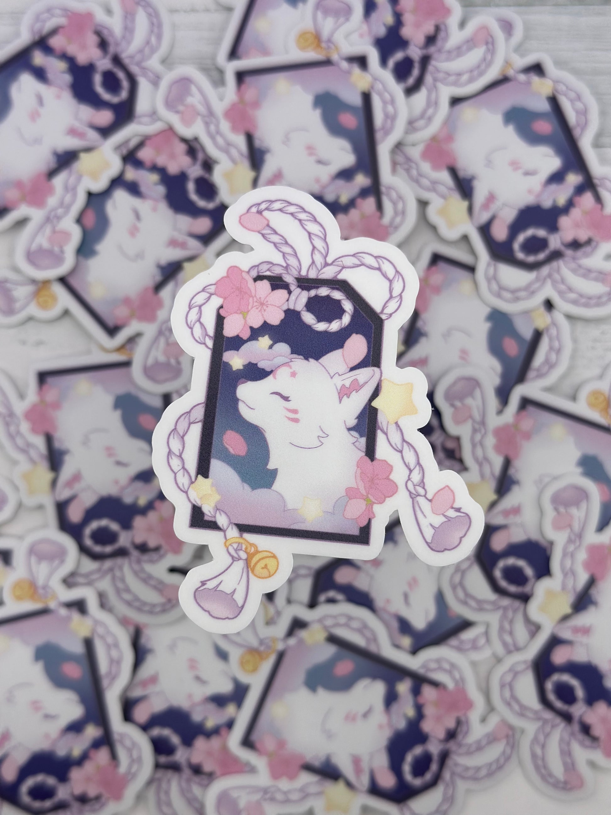 Pastel kitsune sticker framed inside a decorative tag with braided cords, bells, cherry blossoms, and floating stars