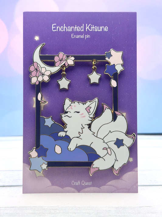 White enchanted kitsune enamel pin resting on blue clouds inside a dark blue frame with cherry blossoms, crescent moon, and dangling star charms on a purple backing card