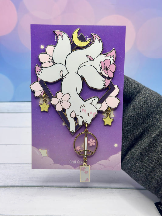 White sakura kitsune enamel pin hanging upside down from a geometric frame with cherry blossoms, crescent moon, dangling stars, and pink lantern charm on a purple backing card