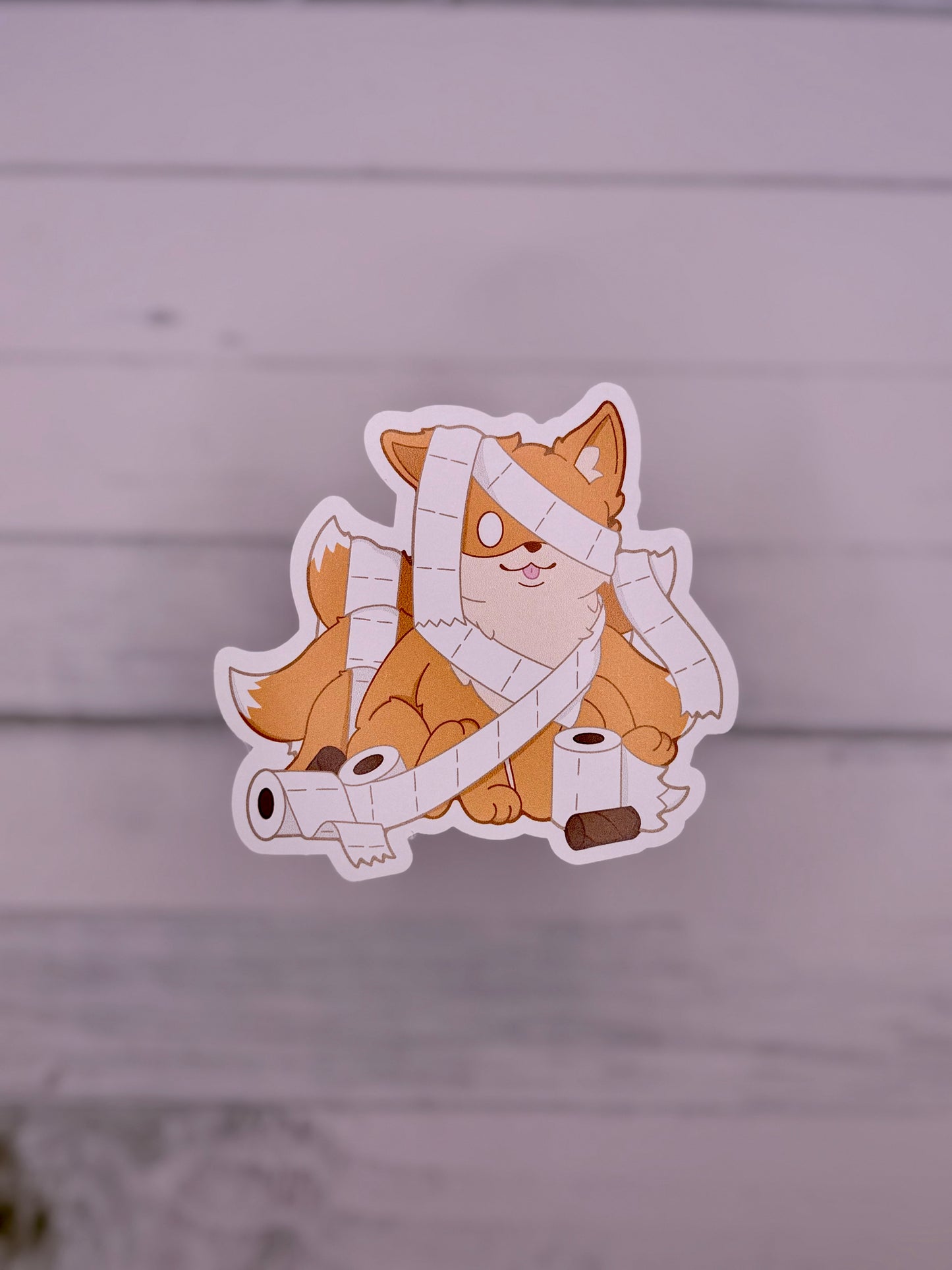 Cute orange kitsune sticker wrapped in toilet paper like a mummy with rolls of toilet paper scattered around it.