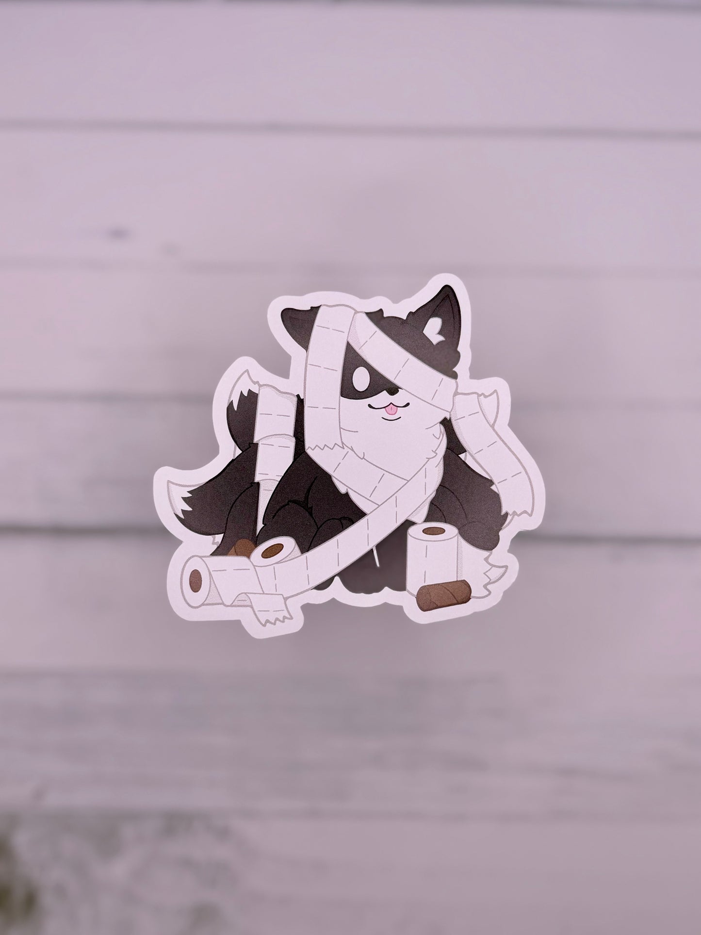 Black kitsune sticker wrapped in toilet paper like a playful mummy surrounded by toilet paper rolls