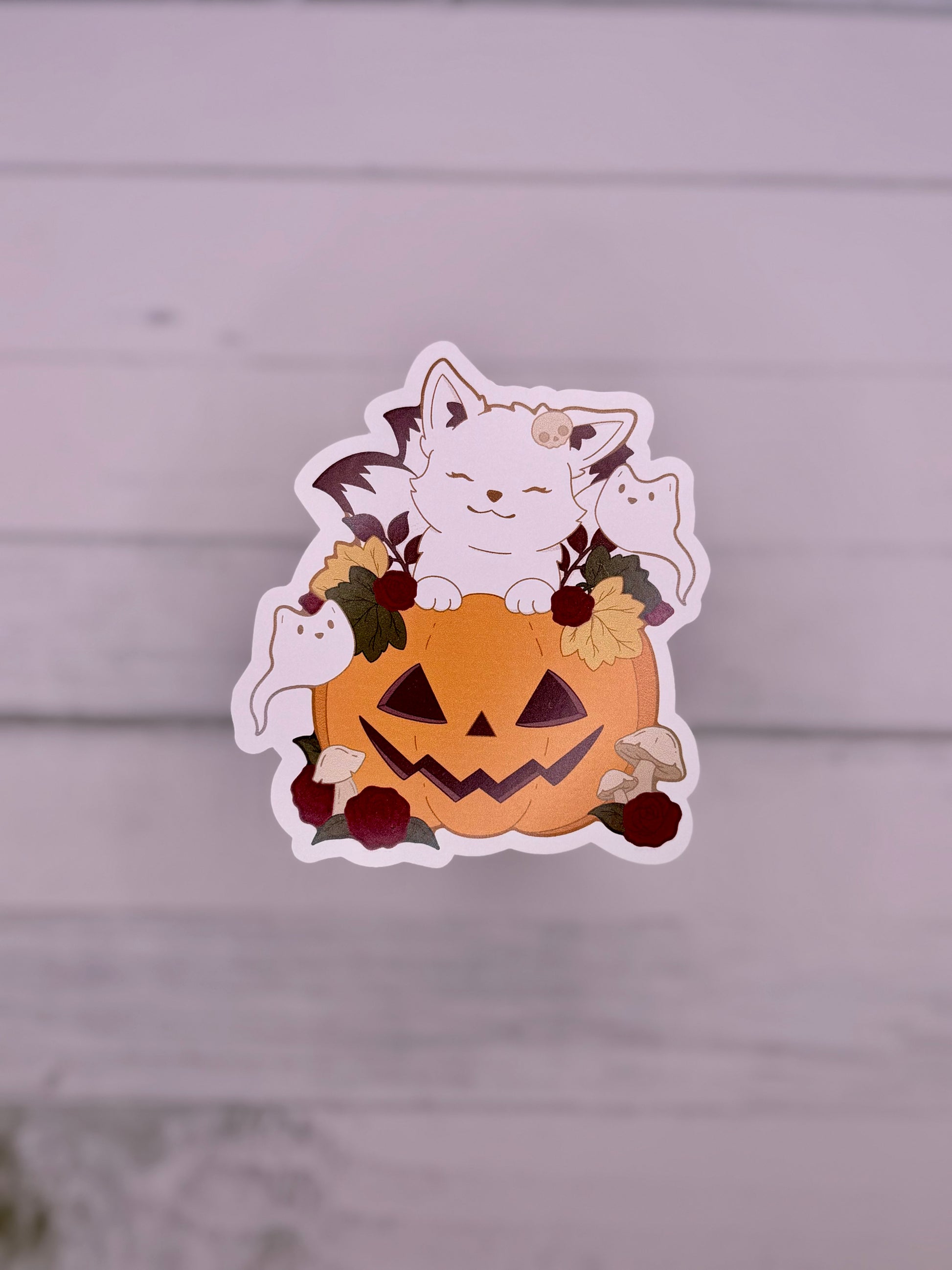 White kitsune sticker resting inside a smiling jack-o-lantern surrounded by flowers, mushrooms, and tiny ghosts