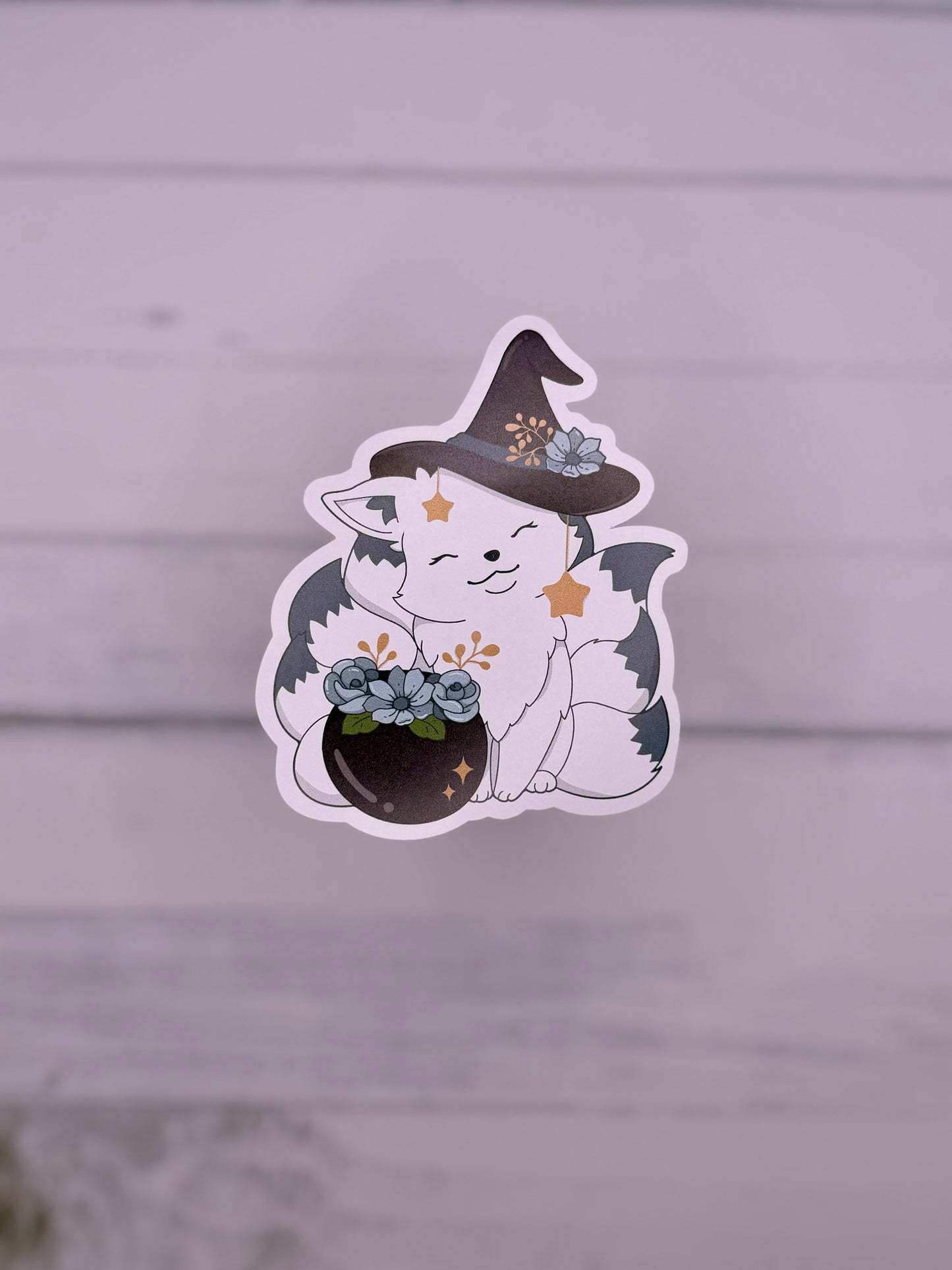 Cute white kitsune sticker wearing a witch hat beside a flower filled cauldron with hanging star charms