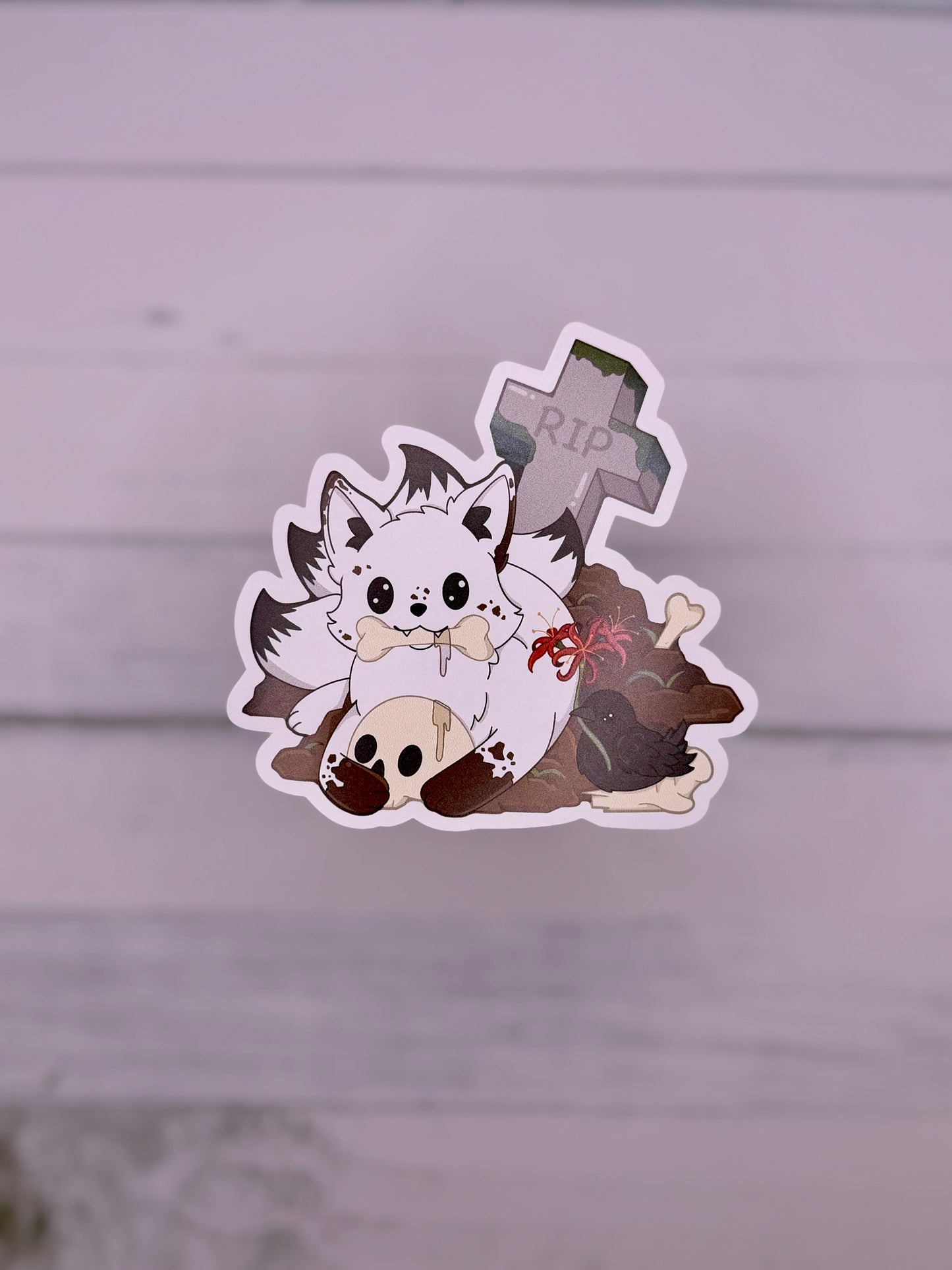 Cute white kitsune sticker sitting in a graveyard with bones, skulls, flowers, and a tombstone