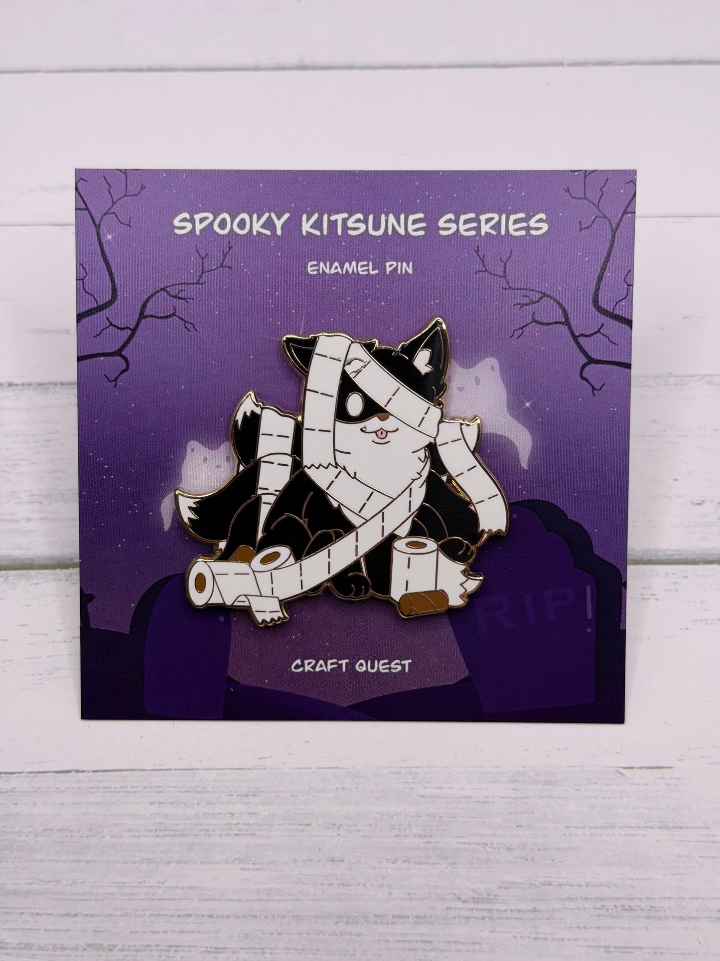 Black and white mummy kitsune enamel pin wrapped in toilet paper with loose rolls scattered around on a purple spooky graveyard backing card