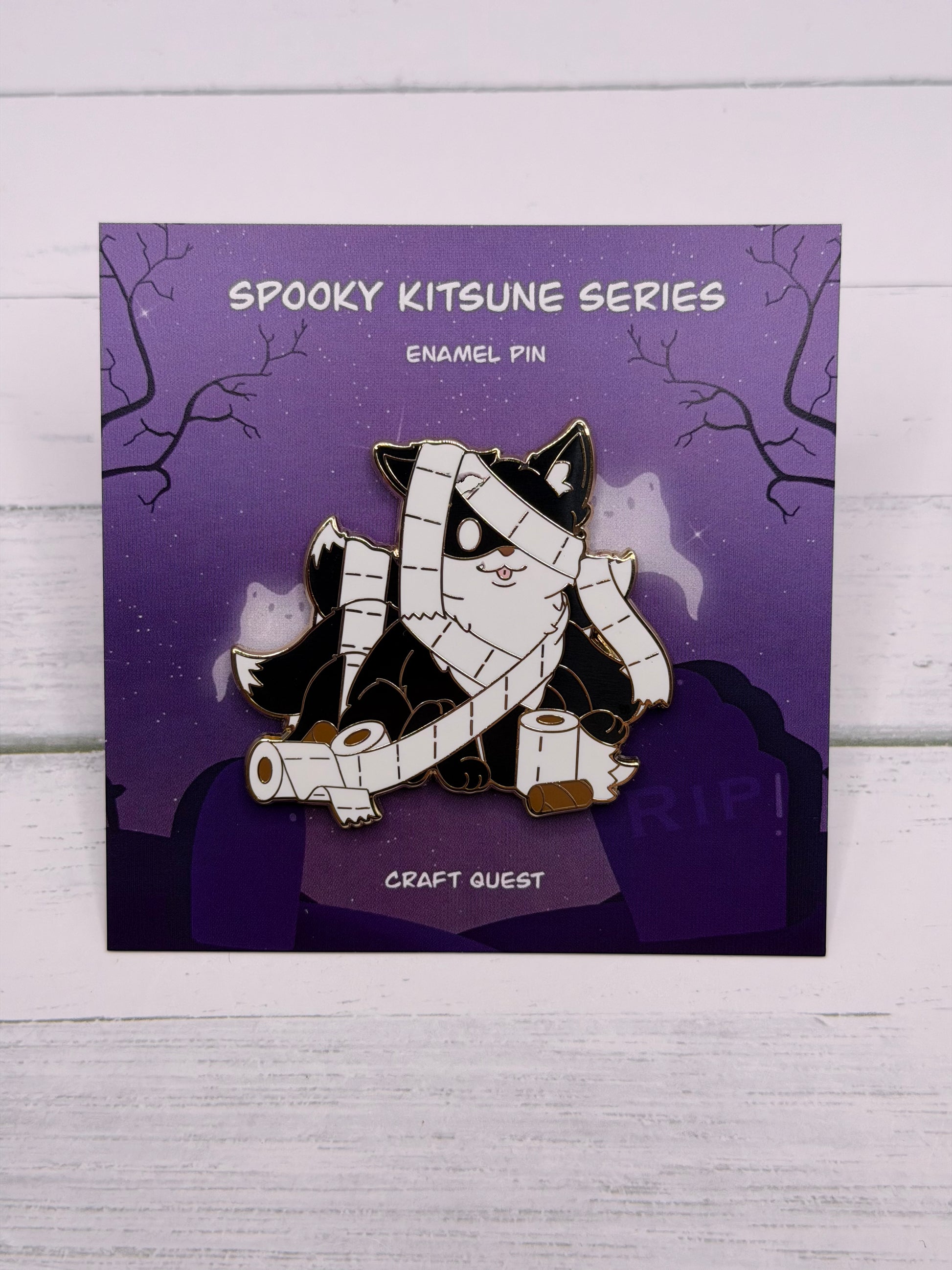 Black and white mummy kitsune enamel pin wrapped in toilet paper with loose rolls scattered around on a purple spooky graveyard backing card