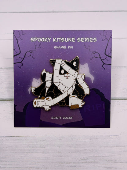 Black and white mummy kitsune enamel pin wrapped in toilet paper with loose rolls scattered around on a purple spooky graveyard backing card