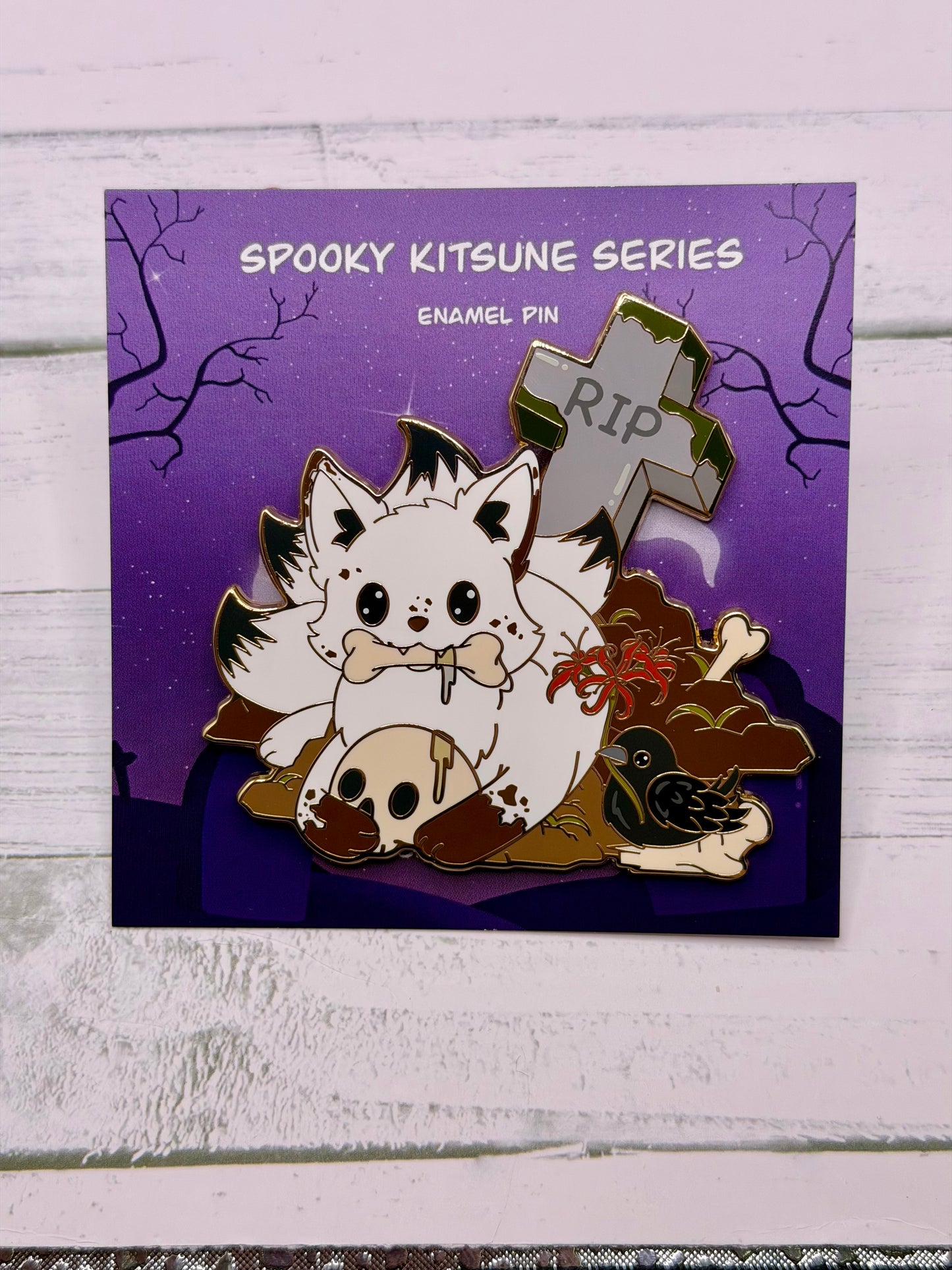 White graveyard kitsune enamel pin holding a bone with a skull, crow, spider lilies, and tombstone details on a purple spooky graveyard backing card