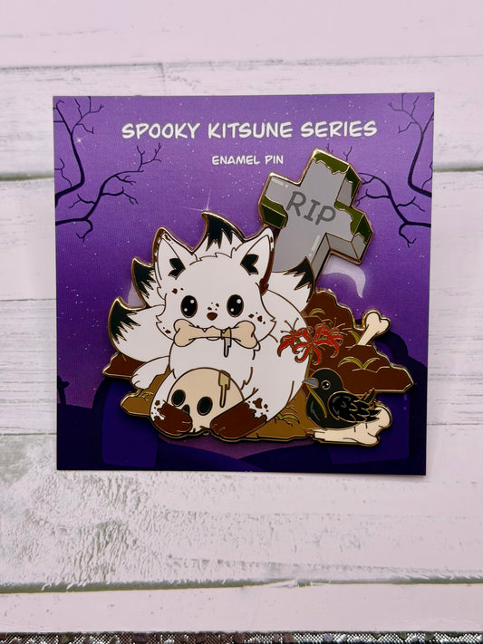 White graveyard kitsune enamel pin holding a bone with a skull, crow, spider lilies, and tombstone details on a purple spooky graveyard backing card