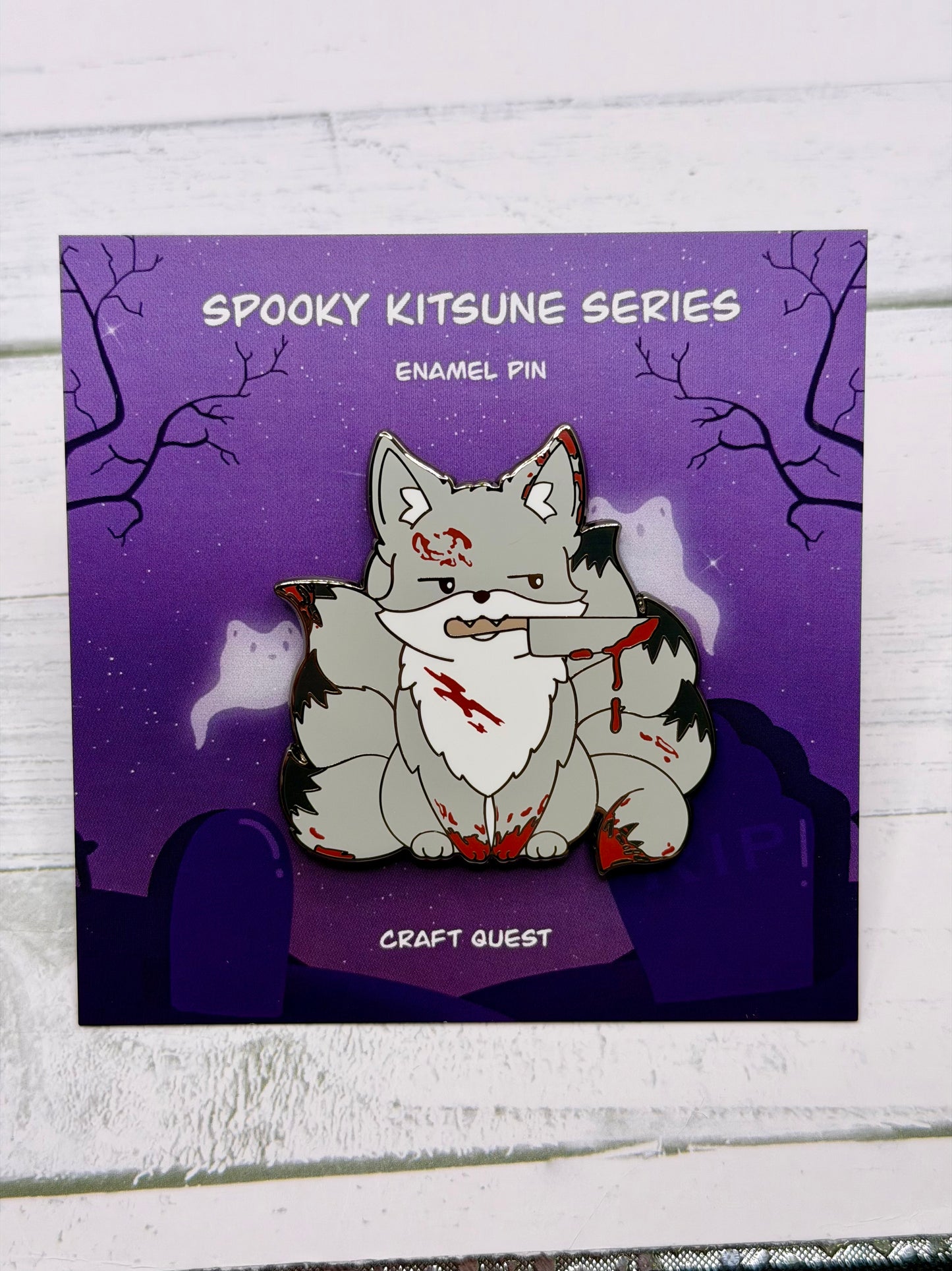 Grey horror themed kitsune enamel pin with blood splatters and a knife held in its mouth displayed on a purple spooky graveyard backing card