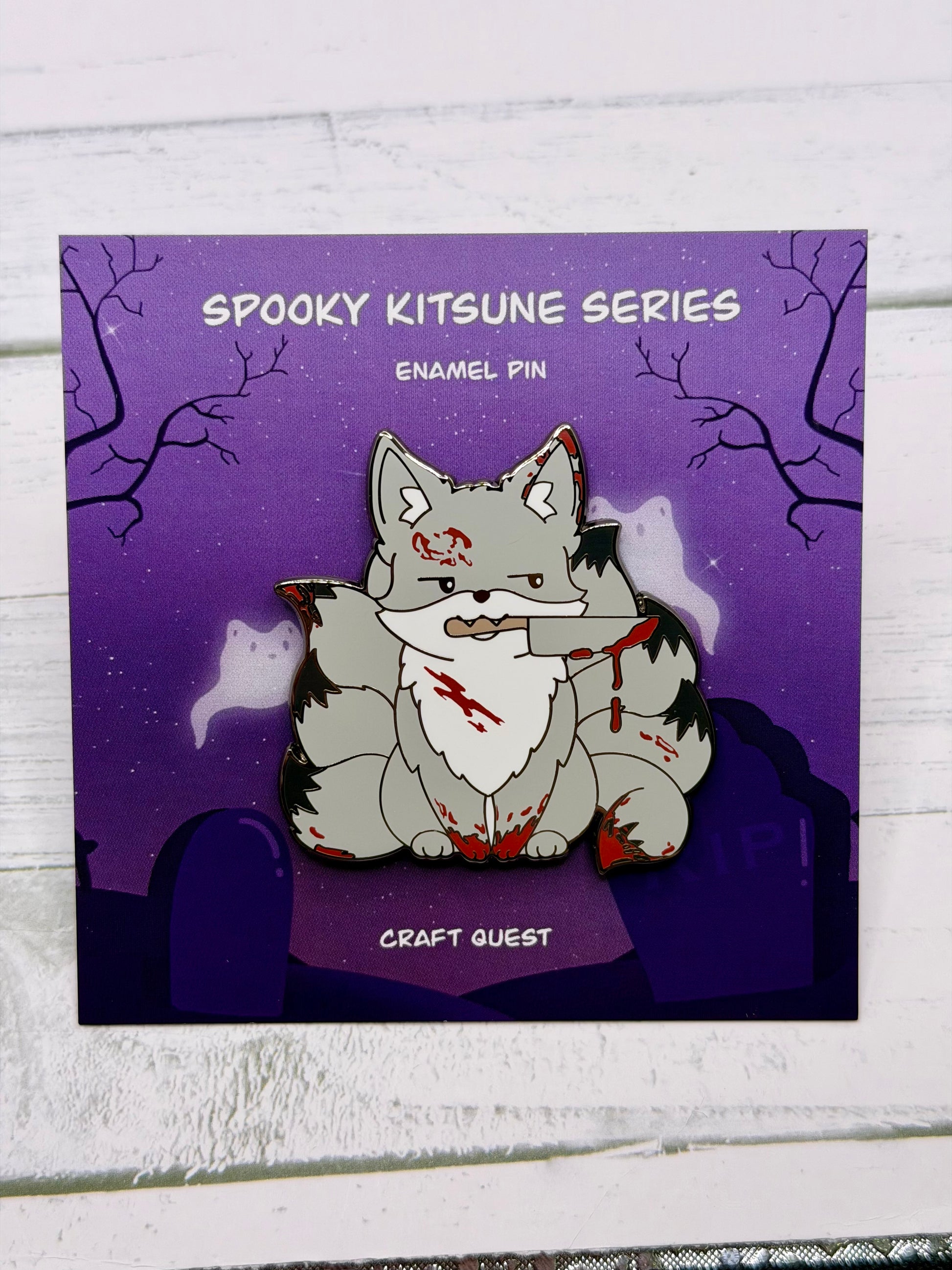 Grey horror themed kitsune enamel pin with blood splatters and a knife held in its mouth displayed on a purple spooky graveyard backing card