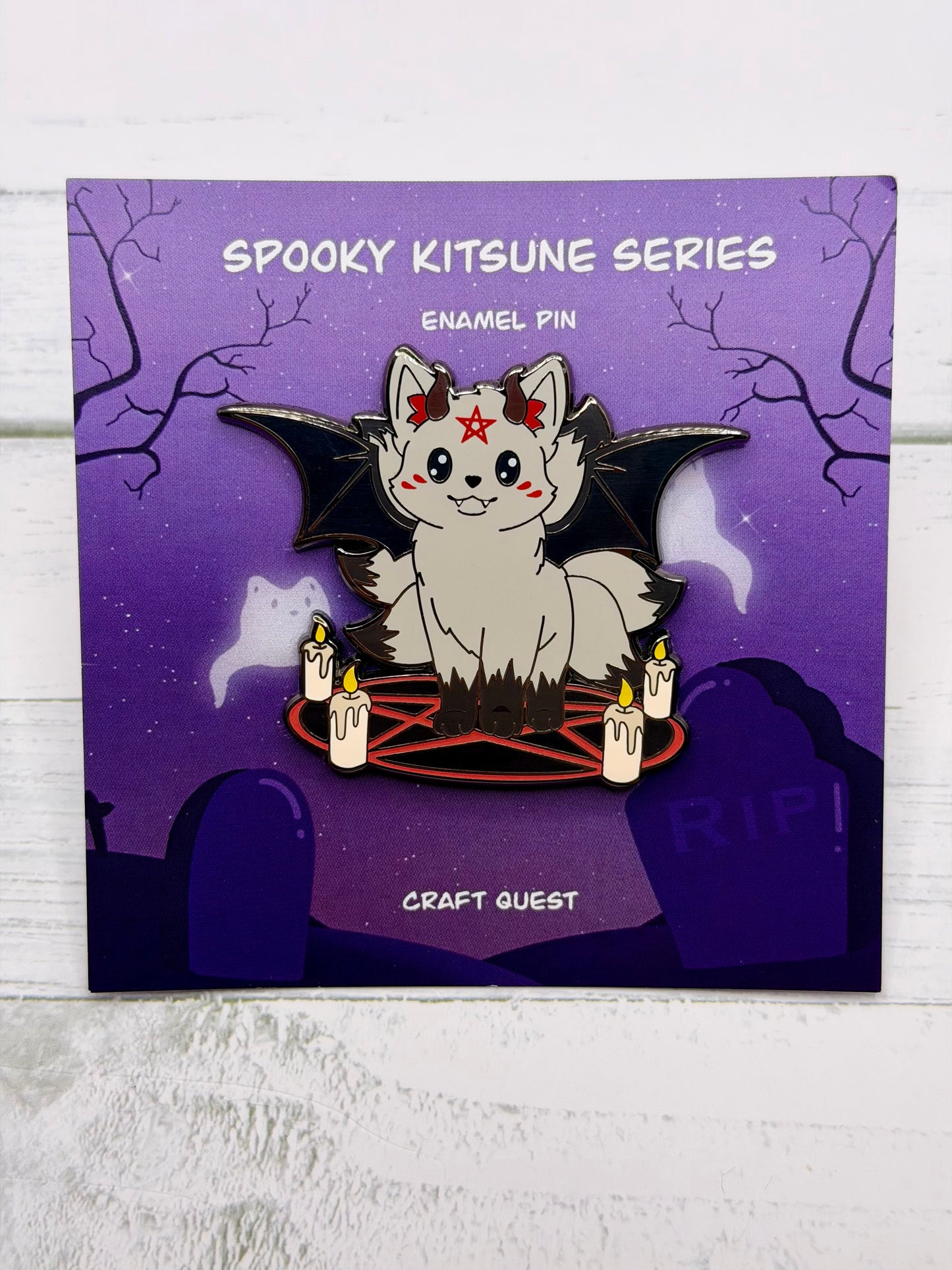 Black kitsune enamel pin with black bat wings, red pentagram circle, candles, and small demon horns in an alternate gold metal version on a purple spooky graveyard backing card.