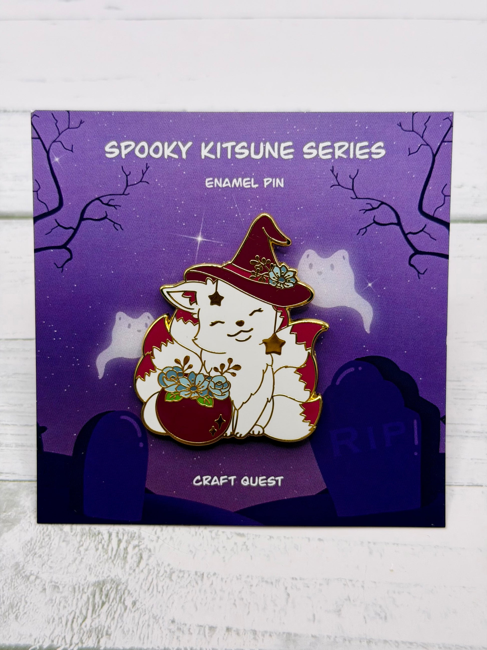 White witch kitsune enamel pin wearing a red witch hat beside a red cauldron filled with blue flowers and gold star charms on a purple spooky graveyard backing card
