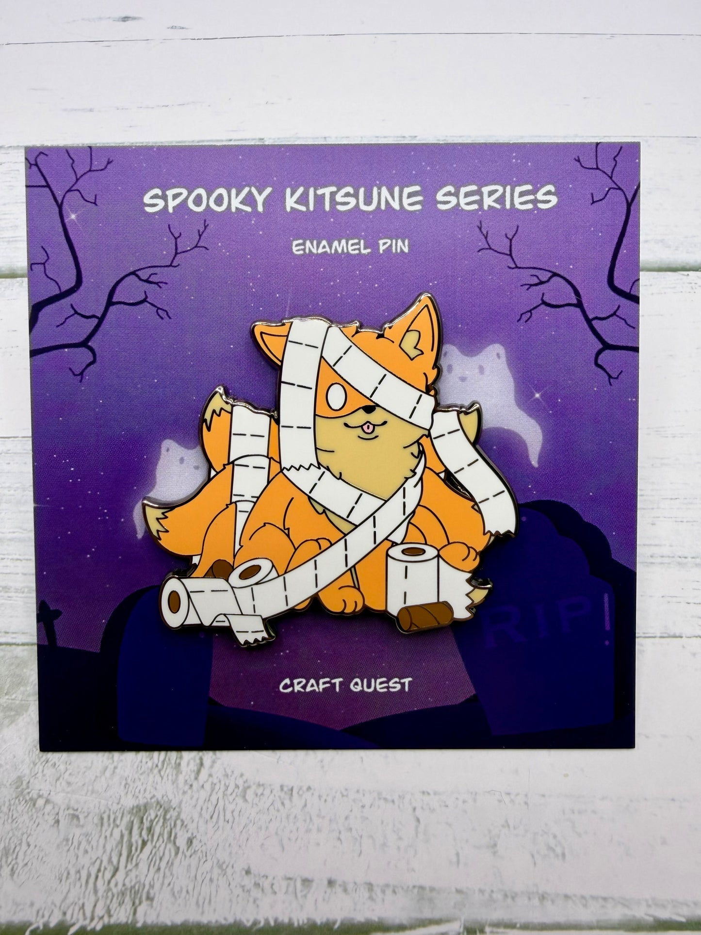 Orange fox mummy kitsune enamel pin wrapped in toilet paper with loose rolls scattered around on a purple spooky graveyard backing card