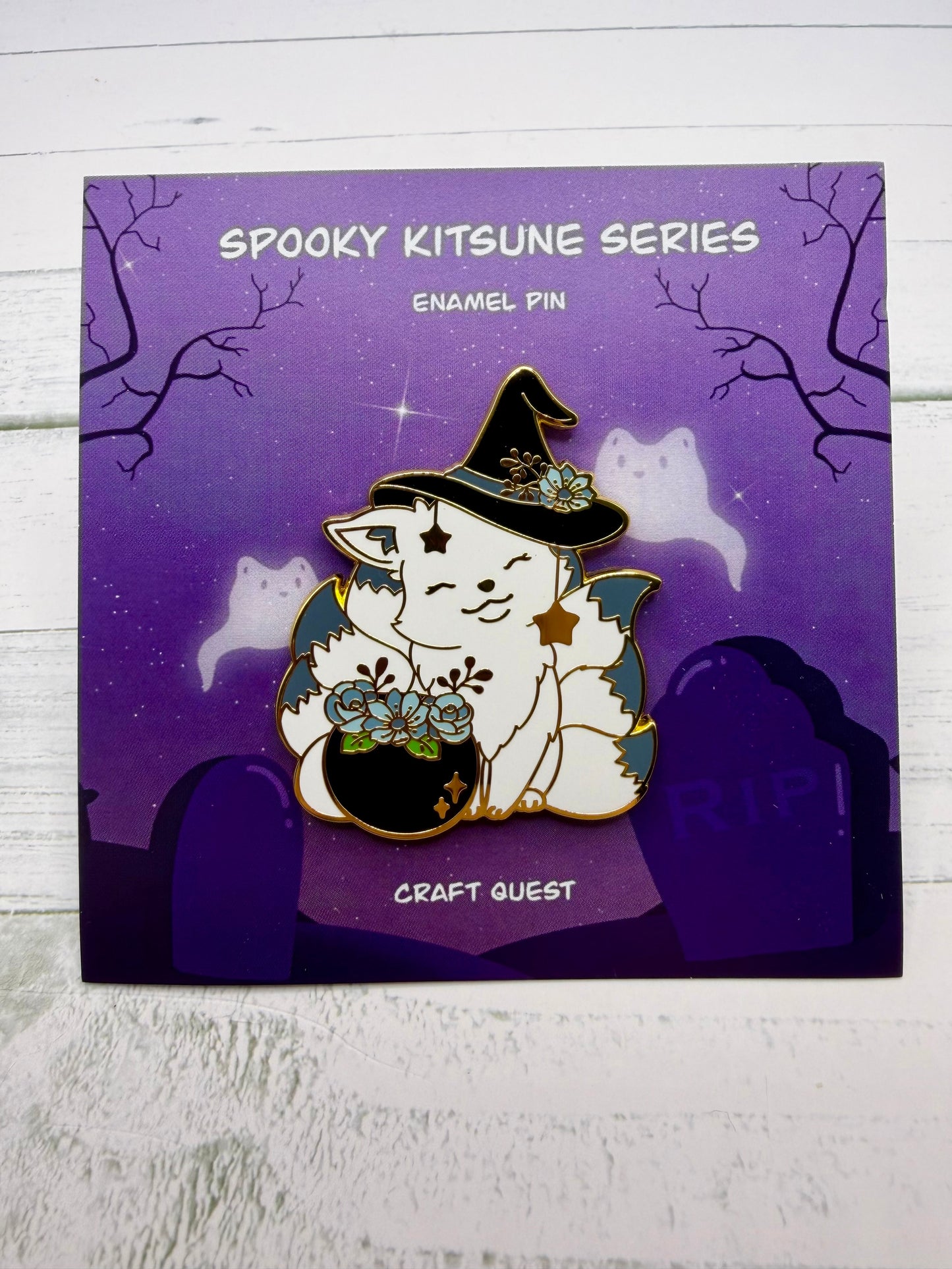 White witch kitsune enamel pin wearing a black hat beside a cauldron filled with blue flowers and gold star charms on a purple spooky graveyard backing card