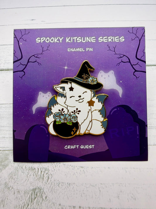 White witch kitsune enamel pin wearing a black hat beside a cauldron filled with blue flowers and gold star charms on a purple spooky graveyard backing card