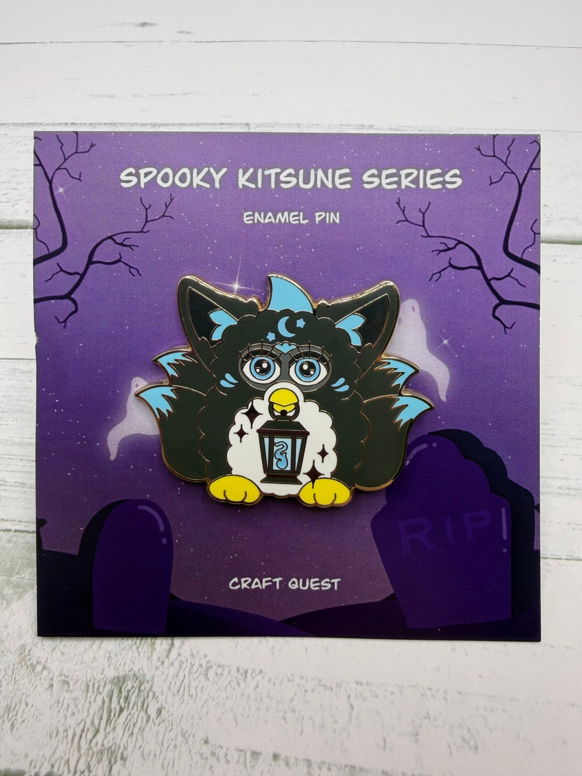 Black and blue retro electronic pet inspired kitsune enamel pin holding a lantern on a purple spooky graveyard backing card