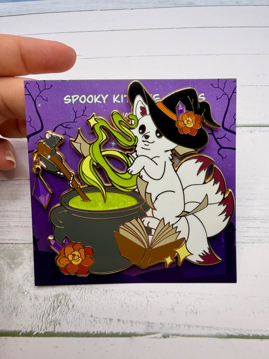 Large white witch kitsune enamel pin stirring a glowing green cauldron with an open spellbook, flowers, purple crystal charm, and dangling rat accessory on a purple spooky graveyard backing card