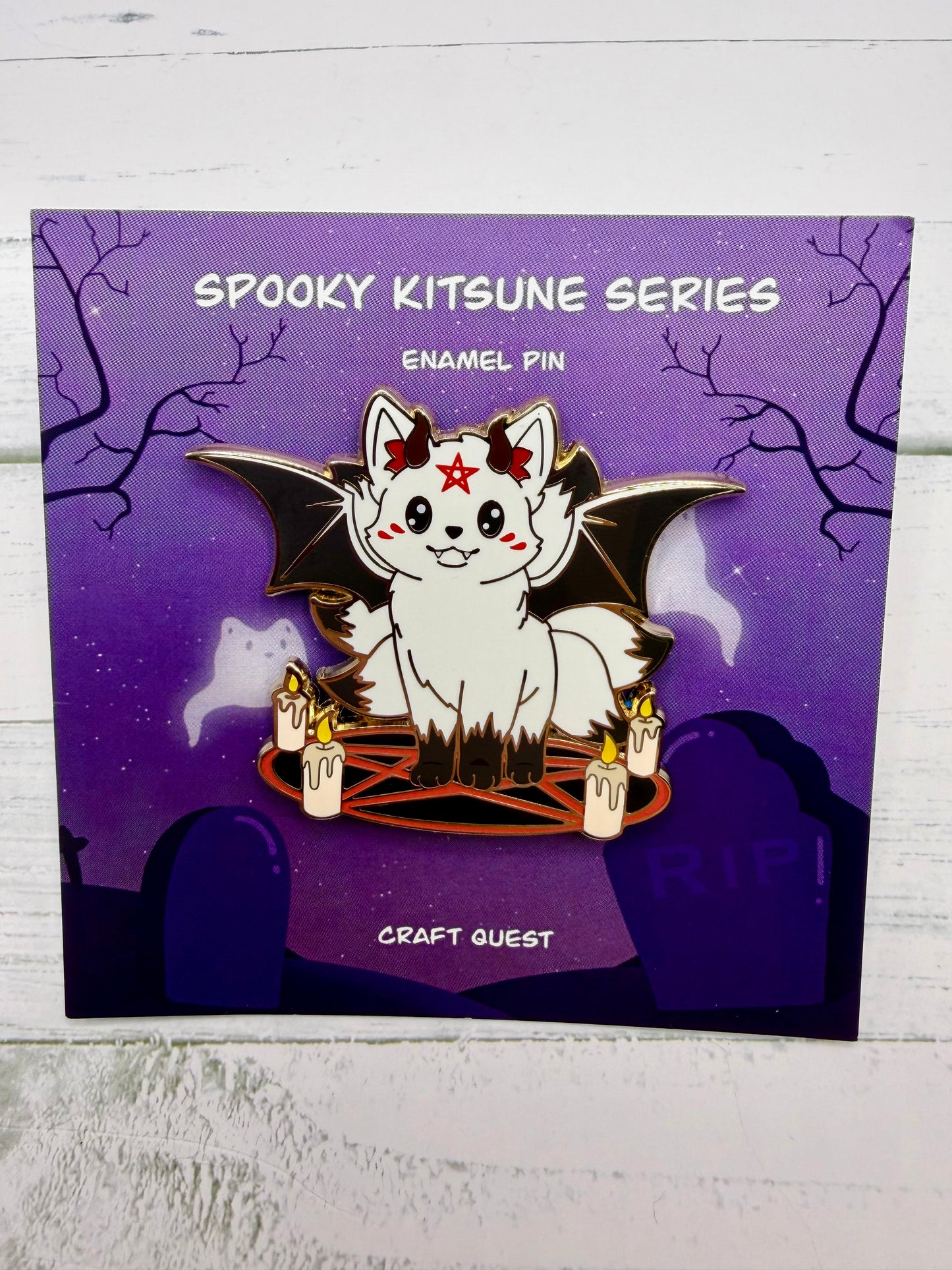 White kitsune enamel pin with black bat wings, red pentagram circle, candles, and small demon horns in an alternate gold metal version on a purple spooky graveyard backing card.