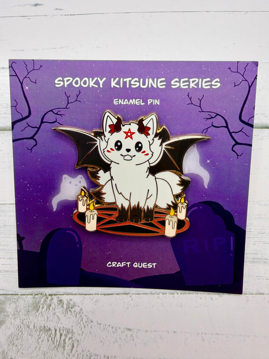 White kitsune enamel pin with black bat wings, red pentagram circle, candles, and small demon horns in an alternate gold metal version on a purple spooky graveyard backing card.