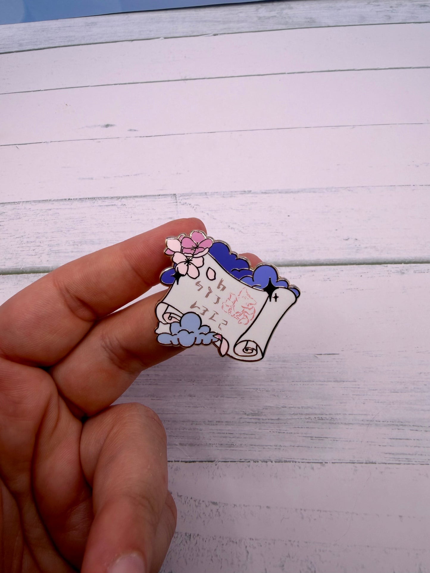 Elegant enamel pin featuring a rolled scroll with Japanese inspired text, sakura flowers and blue clouds