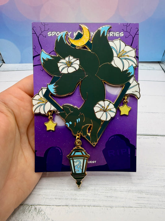Dark enchanted kitsune enamel pin hanging upside down from a geometric frame with white lilies, crescent moon, dangling stars, and blue lantern charm on a purple backing card