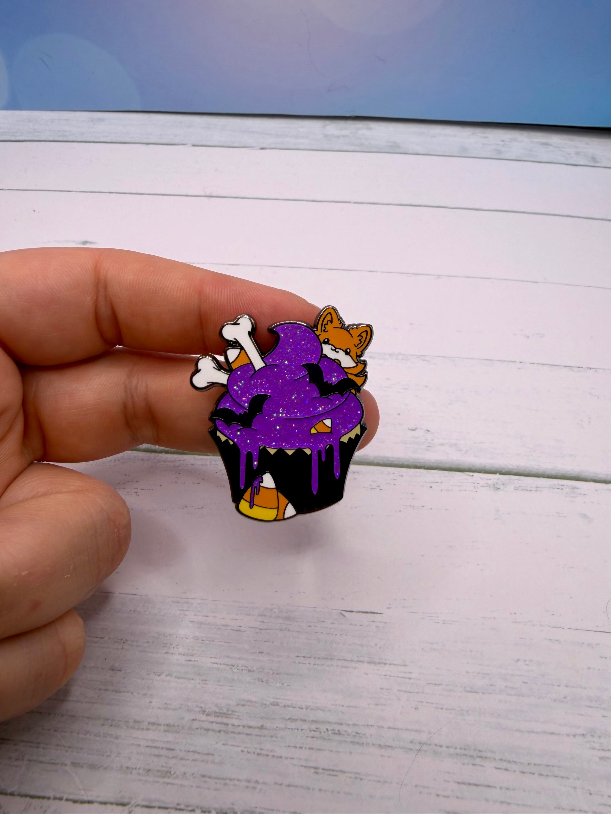 Cute Halloween cupcake enamel pin with glittery purple frosting, candy corn, bat decorations, bones, and a tiny fox peeking out.